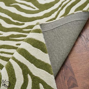 Op de afbeelding: Een getuft karpet met een olijfgroen en crèmekleurig zebrastrepenpatroon. Het kleed is gedeeltelijk omgevouwen om een grijze stoffen achterkant en een getextureerd oppervlak te onthullen. Een klein zwart logo met de tekst KNOTS OVERRIDE is zichtbaar.