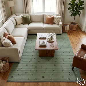 Op de afbeelding: Een woonkamer met een grote, lichtgekleurde hoekbank en diverse kussens. Een houten salontafel staat op een groen tapijt met een geometrisch pijlenpatroon. De kamer is goed verlicht met natuurlijk licht.