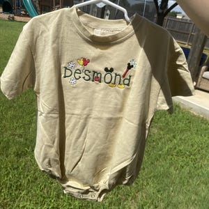 May include: A beige short-sleeve romper featuring the embroidered name "desmond" in dark green letters. The design includes colorful embroidered accents such as a white glove, red and yellow balloons, a black mouse ear silhouette, and a magic wand.