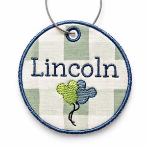 May include: Embroidered round luggage tag with the name "Lincoln" in navy blue. The tag features a green and white gingham pattern, a blue border, and two Mickey Mouse-shaped balloons in green and blue. A silver-colored wire loop is attached.