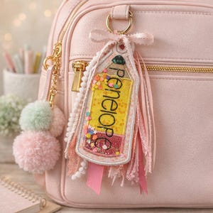 May include: A pink backpack with a gold zipper and a decorative keychain. The keychain is a pencil-shaped tag with the name "Penelope" in yellow, with colorful confetti and pink ribbons. A pink pom-pom keychain hangs on the side.