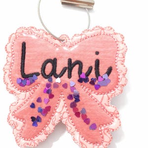 May include: A pink bow-shaped name tag embroidered with the name "Lani" in black. It features heart-shaped sequins in purple and white. A silver clip and wire are attached to the top. The tag is a decorative item.