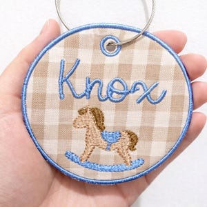 Op de afbeelding: Geborduurde ronde tag met de naam "Knox" in blauw script. De tag heeft een bruin en wit geruit patroon en een blauwe rand. Een schommelpaardontwerp is onder de naam geborduurd. Een zilverkleurige draadlus is aan de bovenkant bevestigd.