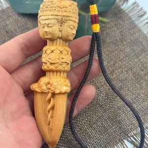 Puede incluir: Colgante de madera tallada de color marrón claro con tres rostros serenos apilados sobre un diseño ornamentado y detallado. El colgante tiene una base puntiaguda y está sujeto a un cordón negro con una cuenta blanca y detalles amarillos y granates.