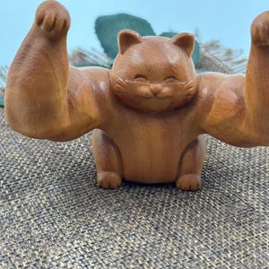 Op de afbeelding: Een houten kattenbeeldje met gespierde armen omhoog in een flex-houding. De kat heeft een lachend gezicht en is gesneden uit lichtbruin hout. Het beeldje is een uniek decoratief item.