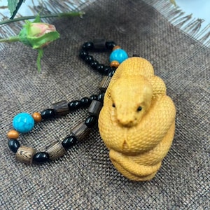 May include: A carved wooden snake figurine in light brown, coiled in on itself. It is accompanied by a beaded bracelet with black, brown, turquoise, and orange beads. The bracelet is attached to the snake.