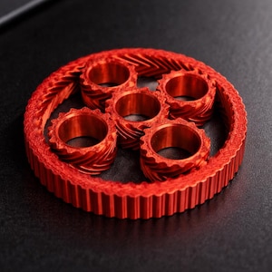 Può includere: Un gruppo di ingranaggi rosso brillante, stampato in 3D, con un anello esterno dentato e cinque ingranaggi circolari interni. Gli ingranaggi sono disposti in un motivo circolare, che mostra un design meccanico. L'immagine è su uno sfondo scuro.