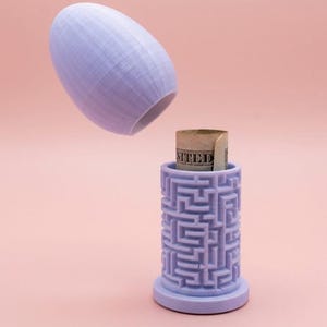 May include: A light purple, 3D-printed puzzle container with a cylindrical base and a removable, egg-shaped lid. The container is open, revealing a rolled-up bill inside. The container has a maze design.