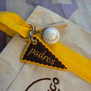 May include: A beige canvas tote bag with a bright yellow strap features a metal keychain with a miniature wooden baseball bat, a white baseball with red stitching, and a brown triangular embroidered pennant with the word padres in yellow.