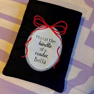 May include: A black fabric Kindle sleeve featuring a central oval patch with a silver glitter finish. The patch is bordered by white lace and topped with a red embroidered bow. The text reads: this is the kindle of a reader, Bella.