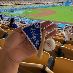 May include: A keyring featuring a blue triangular felt pennant with white scalloped edges and the word dodgers embroidered in white script, attached to a miniature wooden baseball bat and a small white baseball with red stitching.