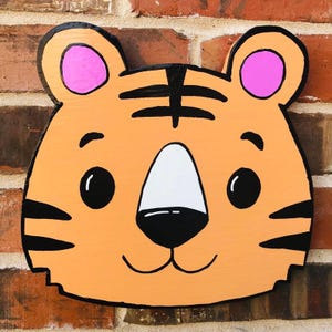May include: A painted wooden tiger head, featuring orange, black, white, and pink colours. The tiger has black stripes, a white nose, and pink inner ears. The cutout is mounted on a brick wall.