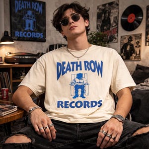 May include: Cream-colored t-shirt with the text "DEATH ROW RECORDS" in blue, along with a blue graphic. The shirt is worn with ripped black jeans and accessories. A poster with the same logo is visible in the background.