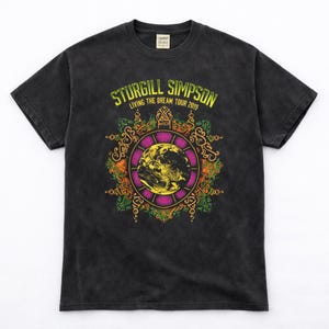 May include: A black Comfort Colors t-shirt featuring the text "STURGILL SIMPSON LIVING THE DREAM TOUR 2019" in yellow. The shirt has a graphic of a globe inside a purple and orange circular design.