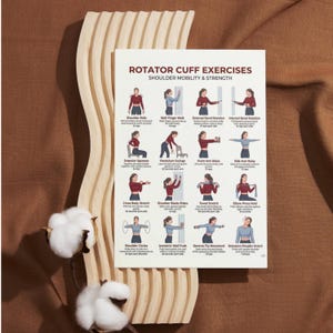 May include: A poster titled "ROTATOR CUFF EXERCISES" with illustrations of shoulder exercises. The poster is on a light wood textured surface, with cotton branches in the foreground. The exercises are for shoulder mobility and strength.