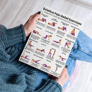 May include: A book titled "Sciatica Pain Relief Exercises" is held by a person. The cover is white with purple text and illustrations of various stretches. The exercises are designed for adults experiencing sciatic nerve pain.