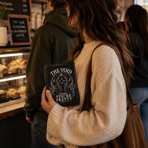 Könnte beinhalten: Eine Person hält ein schwarzes Buch mit dem Text "THE VOID" und "THINKS YOU'RE PRETTY" in Weiß. Das Buch zeigt eine himmlische Illustration. Die Person trägt einen cremefarbenen Pullover und eine braune Tragetasche. Eine Kaffeehaustheke ist im Hintergrund zu sehen.