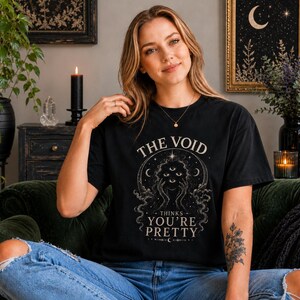 Könnte beinhalten: Schwarzes T-Shirt mit dem Text "THE VOID THINKS YOU'RE PRETTY" in einem kreisförmigen Design. Das Design beinhaltet eine Himmelsillustration mit Mond und Sternen. Die Person trägt eine blaue Jeans.