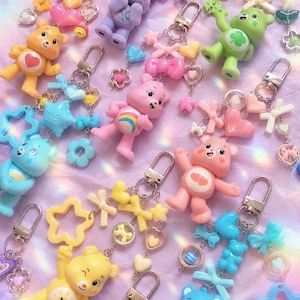 Könnte beinhalten: Eine Sammlung bunter bärenförmiger Schlüsselanhänger mit verschiedenen pastellfarbenen Figuren, die Symbole auf ihren Bäuchen tragen. Jeder Schlüsselanhänger enthält silberfarbene Metallverschlüsse, kleine Kunststoffschleifen, Herzen, Sterne und blumenförmige Anhänger.