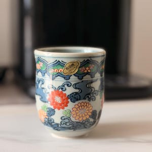 Puede incluir: Una pequeña taza cilíndrica de cerámica con un diseño floral. La taza presenta patrones florales naranjas, azules y verdes sobre un fondo blanco. Una pegatina dorada con texto japonés está adherida al borde. La taza es probablemente para té o sake.