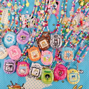 Puede incluir: Una colección de collares de cuentas de colores con colgantes de juguetes digitales para mascotas. Los colgantes presentan varios personajes de dibujos animados en una variedad de colores, incluyendo rosa, azul, verde y naranja. Las cuentas son de colores arcoíris.