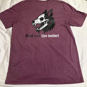 May include: Maroon t-shirt featuring a black and white wolf skull graphic. The text "Dead eyes, Live instinct" is printed below the graphic. The shirt is made of a soft material.