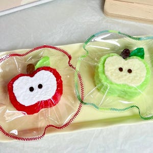 Puede incluir: Una bandeja rectangular amarilla contiene dos jabones con forma de manzana, uno rojo y otro verde, cada uno envuelto en plástico transparente. Los jabones tienen centros blancos y pequeñas hojas verdes.