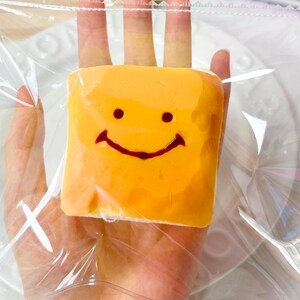 Può includere: Un sacchetto di plastica trasparente che contiene un dolcetto quadrato di colore arancione con una faccina sorridente rossa. Il dolcetto è tenuto in mano, con un calendario di legno e uno sfondo rosa e bianco visibili.