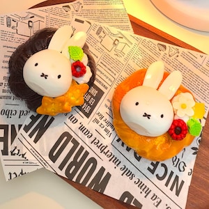 May include: Two decorated donuts with bunny faces, one with a brown glaze and the other with a yellow glaze. Both donuts have white bunny ears and are decorated with flowers and berries.