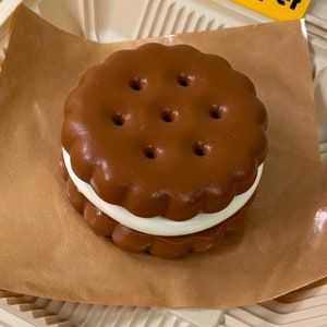 Op de afbeelding: Een chocolade chip cookie sandwich met witte frosting in een bruine, met papier beklede afhaalkarton. De doos heeft een geel sticker met het woord "chipper" erop.
