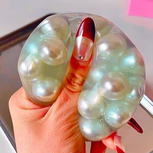 以下が含まれることがあります： 虹色の真珠のような球体で満たされた透明で柔らかなおもちゃ。おもちゃは赤いマニキュアを塗った手で持たれています。