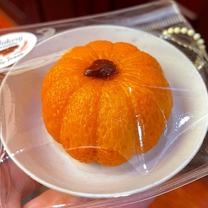 May include: A small orange pumpkin-shaped cake with a brown sugar topping, displayed on a white plate in a clear plastic bag.
