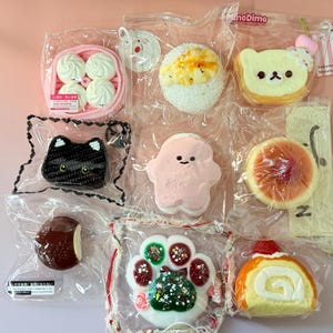 May include: A collection of colorful, packaged squishies in various shapes, including a cat, bear, and dessert items. The items are in clear plastic packaging, and the background is a light pink.