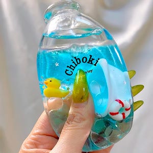 Puede incluir: Una botella transparente llena de líquido azul, con un patito de goma amarillo, un salvavidas blanco y el texto "Chiboki toy". La botella tiene detalles verdes puntiagudos y pequeñas estrellas.
