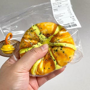 Puede incluir: Primer plano de una mano sosteniendo un pastel en una bolsa de plástico transparente. El pastel es redondo, con un glaseado amarillo y naranja y hierbas verdes. Un pequeño objeto amarillo con un asa naranja es visible. También es visible un recibo con texto.