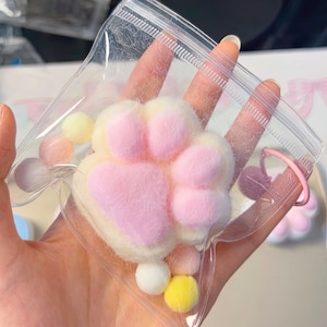 Puede incluir: Un amuleto de pata de gato de color rosa y blanco peludo con un conjunto de pompones coloridos en una bolsa de plástico transparente.