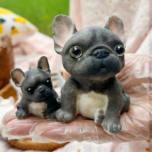 May include: Two detailed, gray French bulldog figurines. The larger one is sitting, and the smaller one is lying down. Both have large, expressive eyes and a white chest. The figurines are likely handmade and are a charming decorative item.