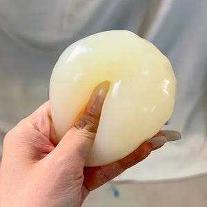 Peut inclure: Une boule translucide, jaune pâle et molle est tenue dans une main. La boule est lisse et semble douce. La main serre légèrement la boule. L'arrière-plan est de couleur claire et neutre.