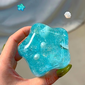 以下が含まれることがあります： 白いパールがあしらわれた、白と銀のグリッターが散りばめられた青い星型のレジン製ペーパーウェイトです。