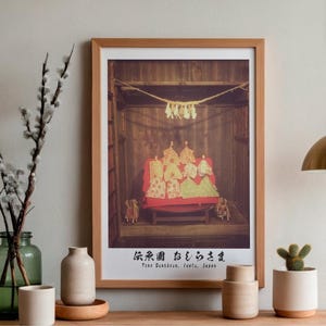 May include: A framed print of a traditional Japanese display featuring several figures in ornate costumes. The print is in a wooden frame and is displayed on a wooden shelf with decorative vases, a small cactus, and a green glass vase with branches.