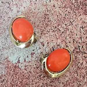 May include: A pair of vintage clip-on earrings. Each earring features a large, oval, coral-colored stone set in a gold-tone metal frame. The earrings are displayed on a textured, reddish-brown surface.