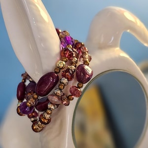 May include: A stack of beaded bracelets in shades of purple, pink, and gold. The bracelets are draped over a white ceramic object with a curved shape. The beads vary in size and shape, creating a textured and colorful accessory.
