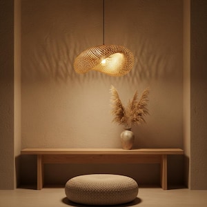 May include: A woven, organic-shaped pendant light fixture hangs above a wooden bench. The light fixture is made of a light brown material and casts a warm glow. A vase with dried plants and a round ottoman complete the scene.