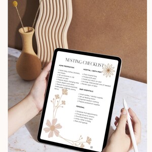Puede incluir: Una tableta digital que muestra una "Lista de verificación de anidación" con encabezados para la preparación del hogar, la preparación del hospital/nacimiento, los elementos esenciales para el bebé y las tareas personales. La tableta se sostiene frente a un fondo neutro.