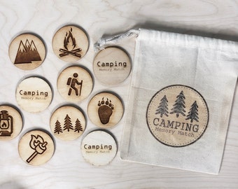 Puede incluir: Un juego de memoria de camping de madera con piezas en una bolsa de cordón blanco. Las piezas presentan varias imágenes con temática de camping, incluyendo una fogata, un excursionista, una huella de pata de oso y una linterna.