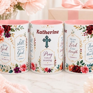 May include: Three white ceramic mugs with pink interiors, decorated with floral patterns in red, pink, and green. The mugs feature religious text including 'Be still, and know that I am God', 'Pray without ceasing', and 'The joy of the Lord is your strength'.
