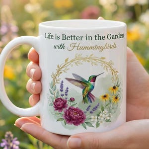 May include: White ceramic mug featuring a colorful hummingbird design with the text "Life is Better in the Garden with Hummingbirds." The mug is decorated with floral elements and a gold-colored wreath. The mug is held in front of a blurred garden background.