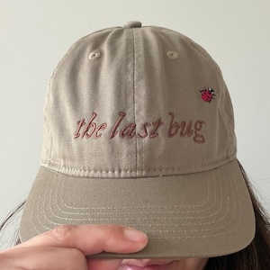 May include: A tan baseball cap with the embroidered text "the last bug" in a reddish-brown thread. A small embroidered ladybug is on the right side of the cap. The cap has a curved brim and a button on top.