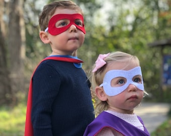 May include: Two children dressed as superheroes. The boy is wearing a red cape and a red felt mask. The girl is wearing a pink and purple cape and a light blue felt mask.