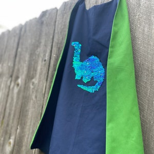 May include: A blue and green reversible cape with a sequined dinosaur design. The dinosaur is green and blue with a shimmery finish.